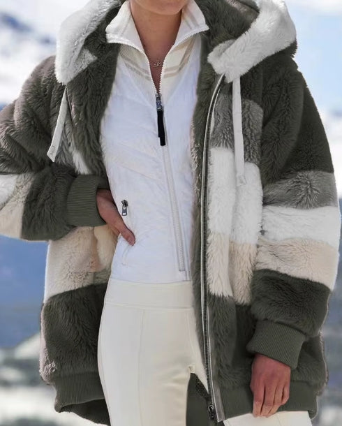 A person wearing a chunky, multi-colored patchwork sherpa fleece jacket with olive green, cream, and white blocks, layered over a white zip-up ski jacket and white ski pants, set against a snowy mountain background.
