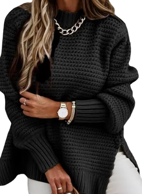 A woman wearing a chunky black knit sweater with a high ribbed mock neck, oversized sleeves, and a side slit, styled with a chunky silver chain necklace, watch, and bracelets.