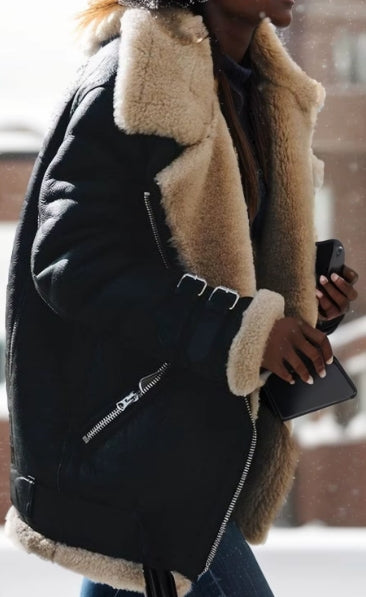 A person wearing a black shearling-lined jacket with a beige sherpa collar and cuffs, holding a black clutch in a snowy outdoor setting.