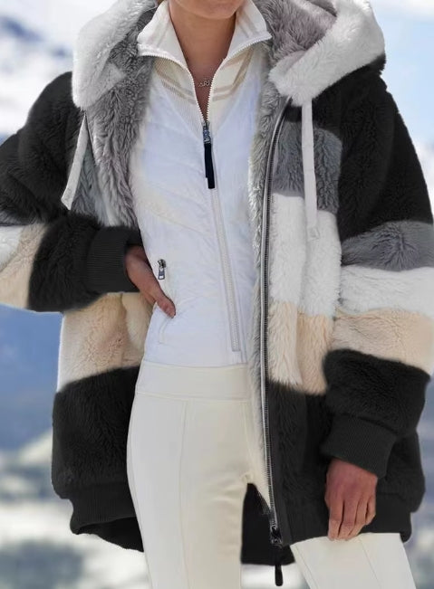 A person wearing a color-blocked sherpa fleece jacket with black, white, gray, and beige panels, layered over a white zip-up ski jacket and white ski pants, set against a snowy mountain background.