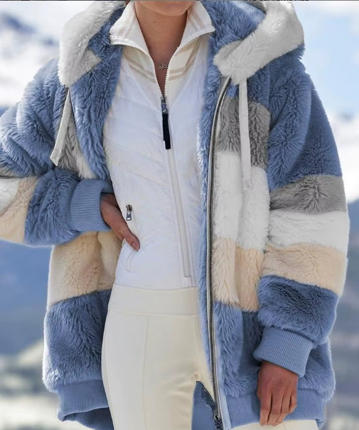 A person wearing a plush color-blocked jacket with blue, cream, beige, and gray panels over a white zip-front top and cream pants, photographed outdoors with a blurred mountain background.