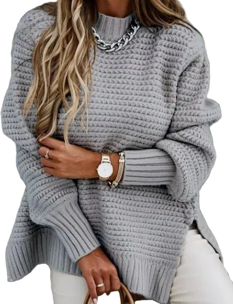 A woman wearing a chunky light gray ribbed knit sweater, styled with a silver chain necklace, gold watch, and bracelets, paired with white pants.