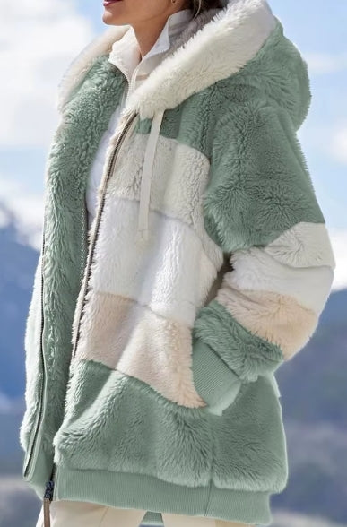 A close-up of a person wearing a plush hooded jacket with soft color-blocked panels in sage green and cream, photographed outdoors against a blurred mountain and sky background.