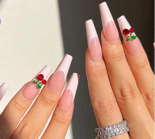 A set of press-on nails shown on a hand, alongside the loose nail tips on a pink background. The nails have a long square tip with a soft pink to white ombre gradient and a small red and green cherry design near the base.