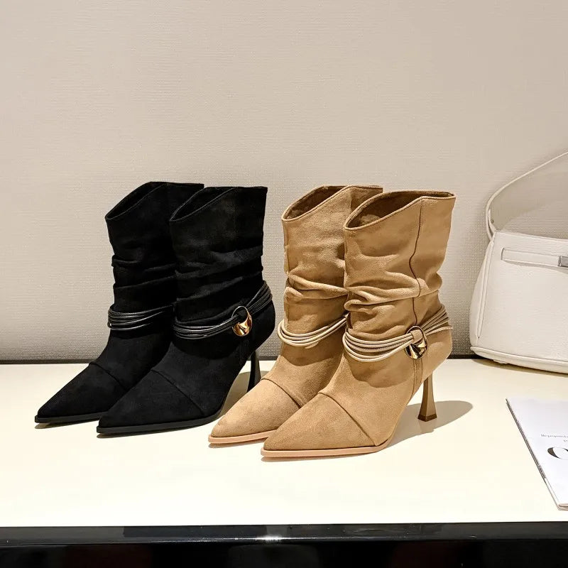 Two pointed-toe mid-calf boots with ruched shafts and kitten heels, shown side by side on a white surface against a light neutral wall. The left boot is black with a metallic gold buckle detail, and the right boot is tan/beige with a white stripe and a small gold buckle detail.