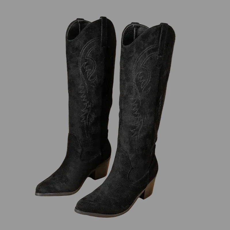 A pair of black suede cowboy boots with pointed toes and low block heels, standing upright on a warm beige background with dried floral decor in the corner.