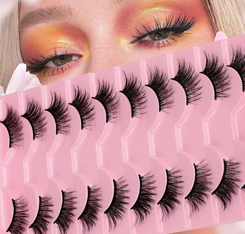 A close-up of a person's eyes wearing dramatic false eyelashes, with a pink tray of multiple pairs of black false eyelashes overlaid across the front. The person has warm orange and gold eyeshadow, and the tray is labeled BY06.