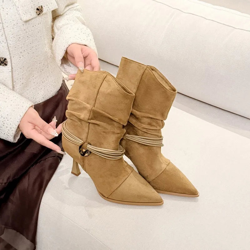 A pair of tan suede ankle boots with pointed toes, stacked ruffle detailing, and a thin metallic strap with a gold buckle around the lower shaft. The boots are held by a person wearing a white textured jacket and a dark brown skirt, seated on a light-colored sofa.