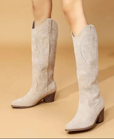 A model wearing knee-high western boots in a light beige suede-like material with embroidered stitching, paired with a brown corduroy mini skirt and a gray cardigan, set against a warm beige studio backdrop.