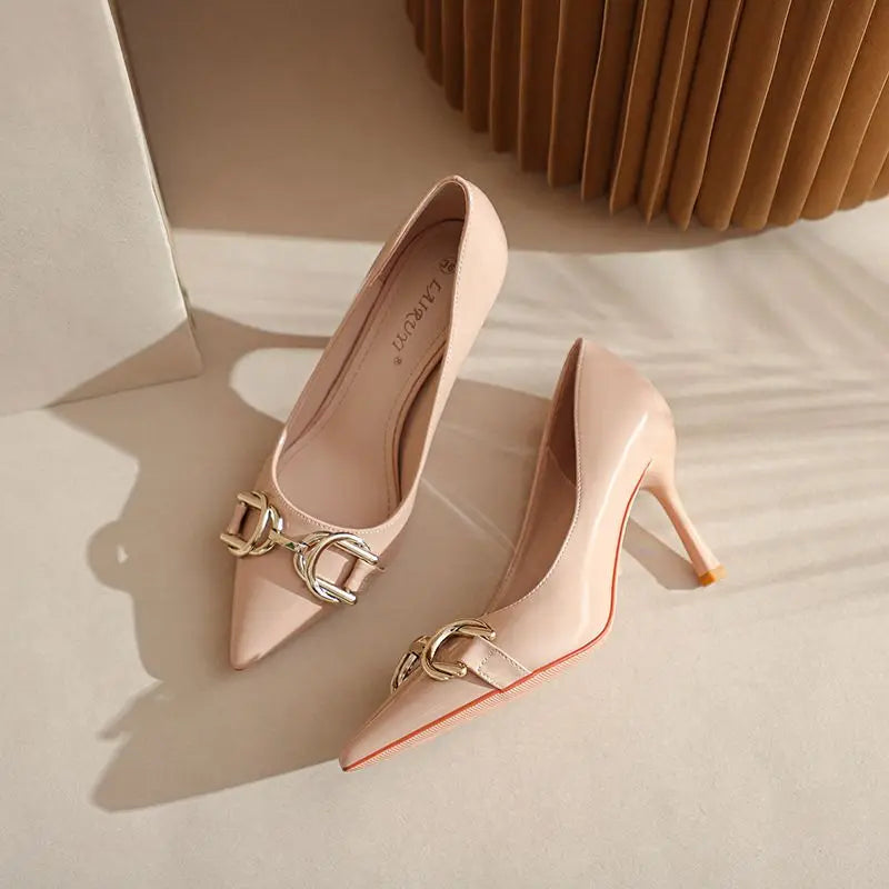 A pair of pointed-toe high heels in a pale blush pink patent leather with gold-tone horsebit hardware, displayed on a beige surface with soft shadows.