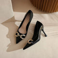 A pair of black pointed-toe kitten heels with a gold-tone horsebit hardware detail across the vamp, displayed on a beige surface with soft shadows.