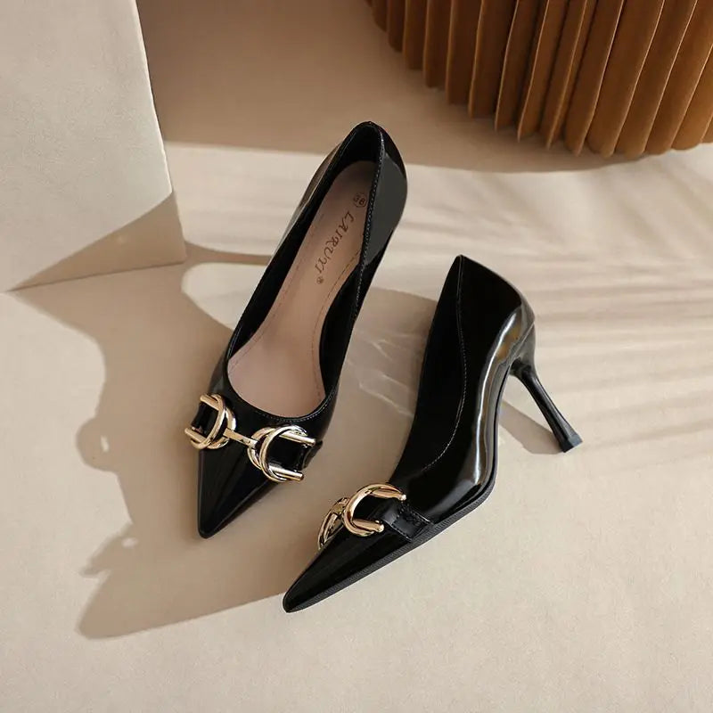 A pair of black pointed-toe kitten heels with a gold-tone horsebit hardware detail across the vamp, displayed on a beige surface with soft shadows.