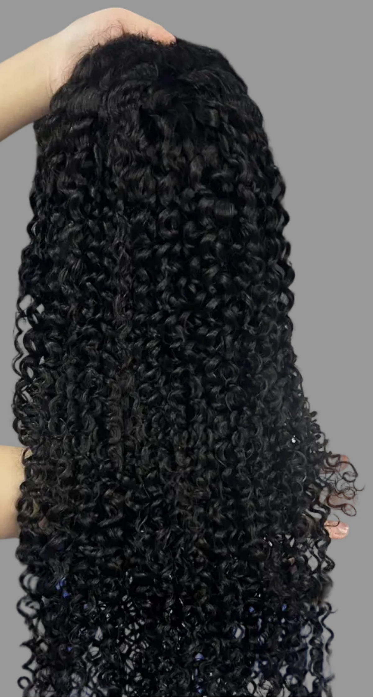 A person holds up a large bundle of long, tightly curled black hair extensions or a wig against a storage rack filled with plastic-wrapped hair pieces. Blue crates and shelving are visible in the background, and the text 'Mona Hair/Kinky Curly' appears at the bottom.
