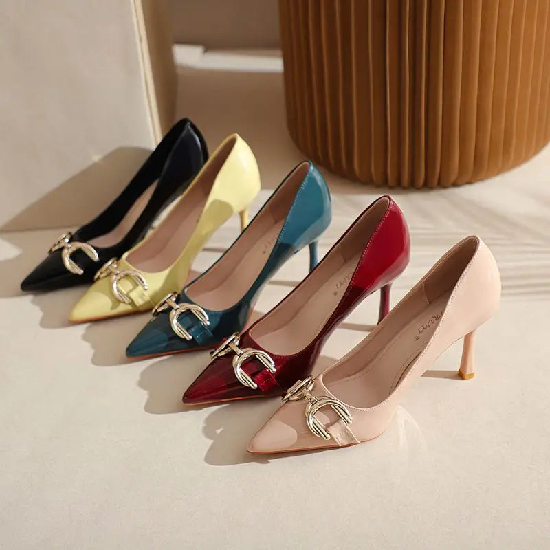 Four pointed-toe high heels arranged diagonally on a light beige surface, featuring shiny patent leather finishes in black, pale yellow, teal blue, burgundy red, and blush pink, each with a large metallic buckle detail on the vamp.