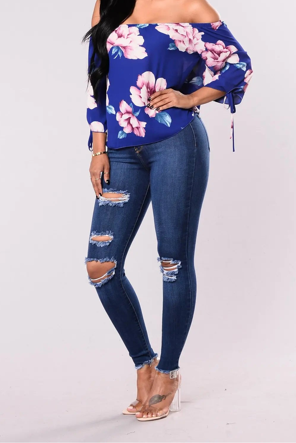 Women's dark blue ripped slim denim leggings styled with a floral off-shoulder top and clear heels, side view
