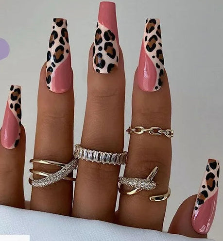 A hand displaying long coffin-shaped nails with pink base and leopard print tips, alongside a small inset showing the nail wrap design pieces. The nails are decorated with multiple gold rings, and the image includes the text '#JP1414' on a lavender banner.