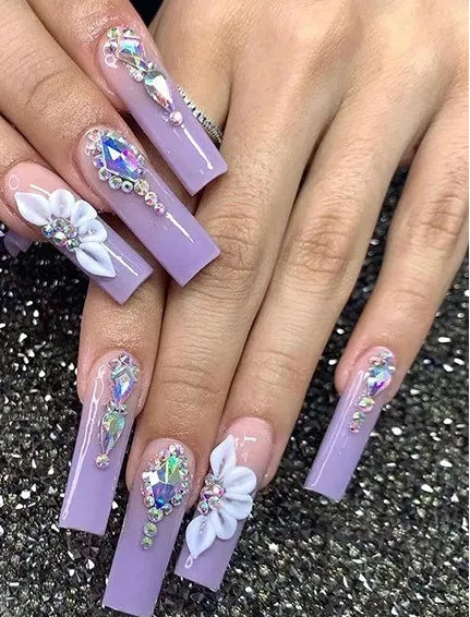 A hand displaying long lavender press-on nails with rhinestone and floral nail art, alongside a small inset showing the individual nail designs and the text '#JP2379'.