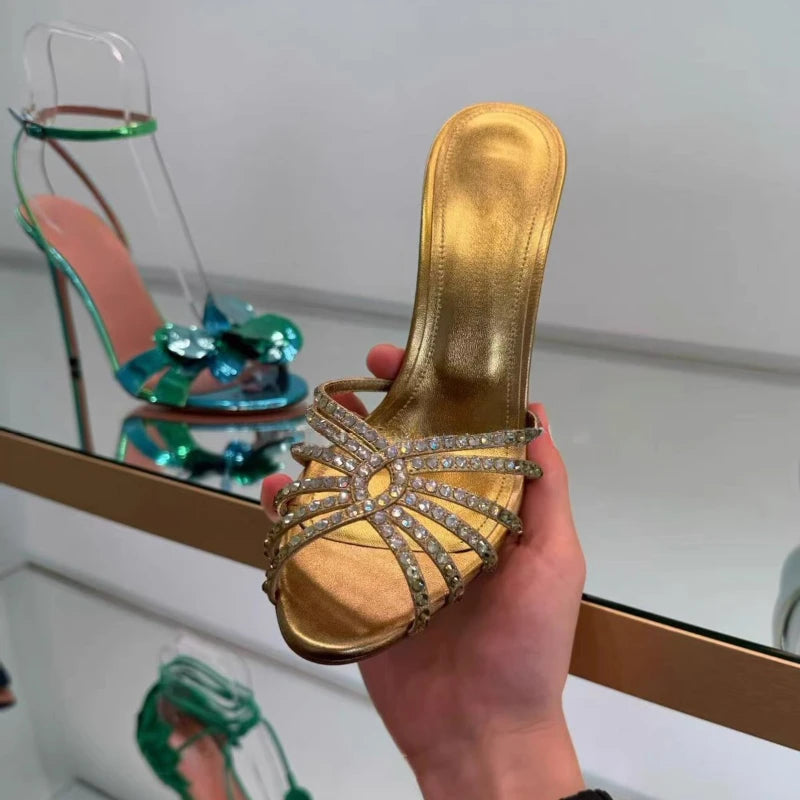 A hand holds a gold open-toe high-heel sandal with crystal-embellished straps. The background shows a mirrored shoe display case with other shoes inside.