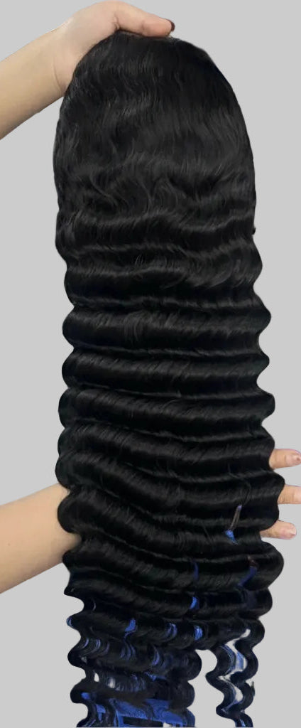 A person holds up a long, straight black wig with deep waves against a plain light gray background. The person is wearing a white knit sweater, and only their hands and partial torso are visible.