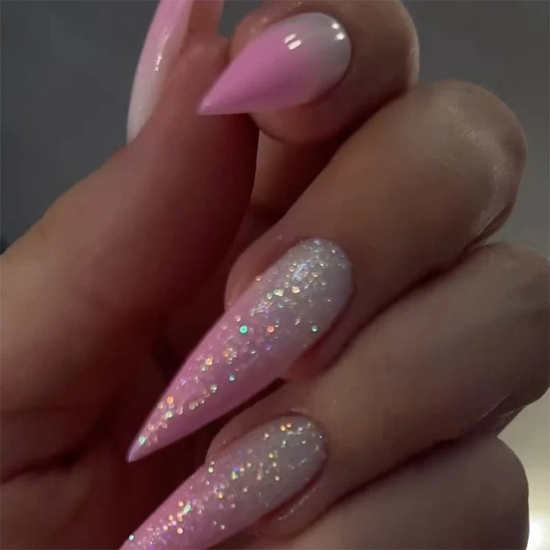 Long stiletto press-on nails in pink-to-nude gradient with iridescent glitter, worn on hand.

