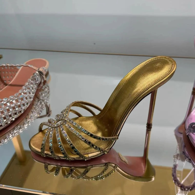 A metallic gold high-heeled mule sandal with crystal-embellished straps, displayed on a reflective gold surface next to other embellished shoes.