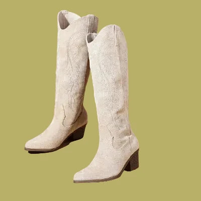 A pair of light beige western-style knee-high boots with pointed toes and low block heels, displayed on a warm beige background with decorative dried grass in the corner.