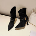 A pair of black pointed-toe ankle boots with a ruched mid-calf shaft, a slim kitten heel, and a decorative metallic ring with a gold heart accent, displayed on a light beige surface next to a white handbag.