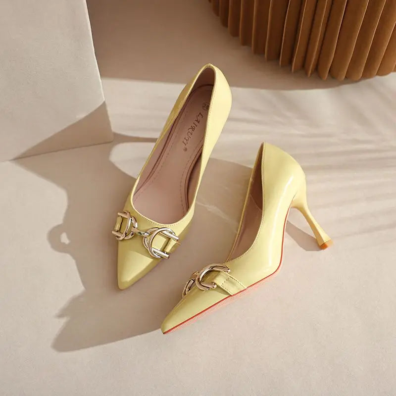 A pair of pale yellow pointed-toe stiletto pumps with a shiny gold-tone chain detail across the vamp and a signature red sole, displayed on a beige surface.