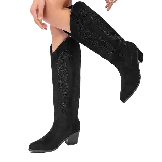 A person wearing black knee-high western-style boots with pointed toes and low block heels, paired with a bright green blazer, photographed against a clean white studio background.