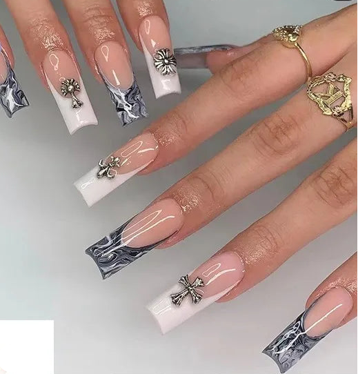 A hand displaying long coffin-shaped press-on nails with a French tip design, some decorated with silver floral and cross charms. The nails feature a gradient from pale pink to white with black marbled detailing at the tips.