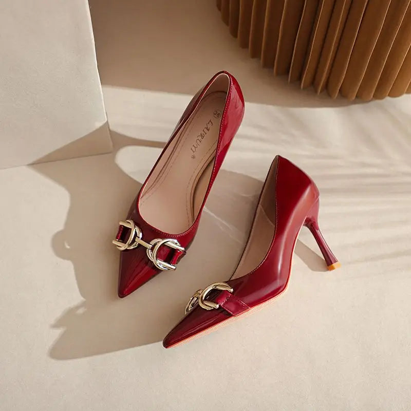 A pair of glossy red pointed-toe kitten heels with a gold-tone horsebit detail on the front, displayed on a beige surface with soft shadows.