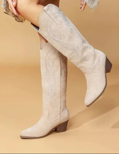 A model wearing a pair of light beige suede knee-high western boots with embroidered stitching, posed against a solid tan studio background.
