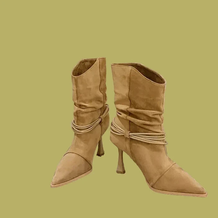A pair of beige suede ankle boots with pointed toes, slim high stiletto heels, and ruched detailing on the shaft, displayed on a textured gray and white checkered surface.