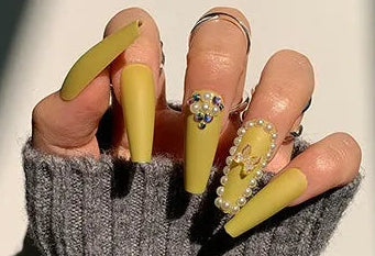 Yellow press-on nails with rhinestone accents shown on a hand wearing a gray knit fingerless glove, alongside a small inset of the individual nail set and the text '# 1312'.