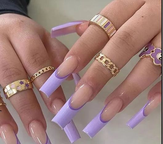 A hand displaying long coffin-shaped press-on nails in a lavender and nude colorway, with a matching nail color swatch inset at the bottom left. The nails feature a French tip gradient transitioning from nude beige to light lavender.