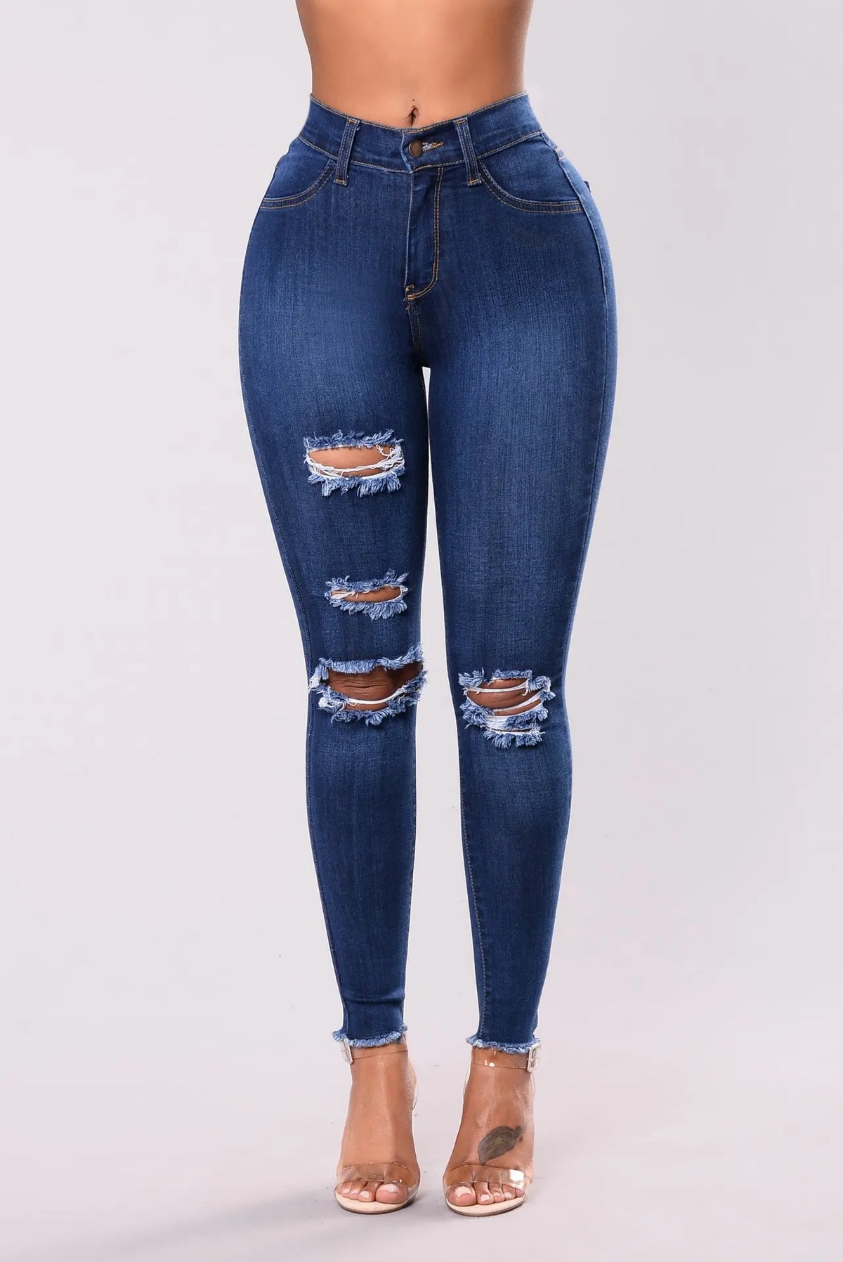 Front view of women's dark blue ripped denim leggings with frayed knee cutouts and raw hem, worn with clear heels
