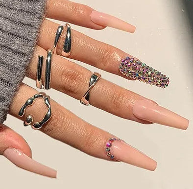 A hand wearing long nude-pink press-on nails with silver rings, alongside a small inset showing the individual nail designs. The main hand is dressed in a gray ribbed sleeve, and the nails are labeled with '#1357'.