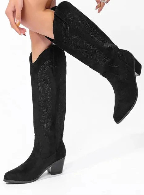 A person wearing black knee-high western-style boots with pointed toes and low block heels, paired with a bright green blazer, photographed against a clean white studio background.