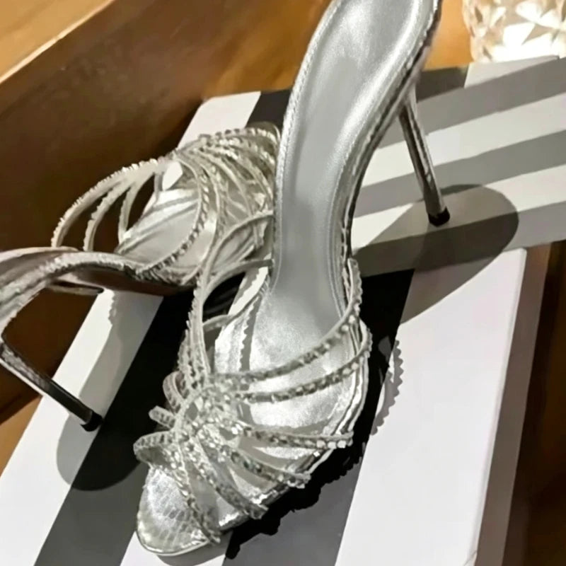 A pair of metallic silver high-heeled sandals with multiple thin straps adorned with small rhinestones, displayed on a white and black striped shoebox.