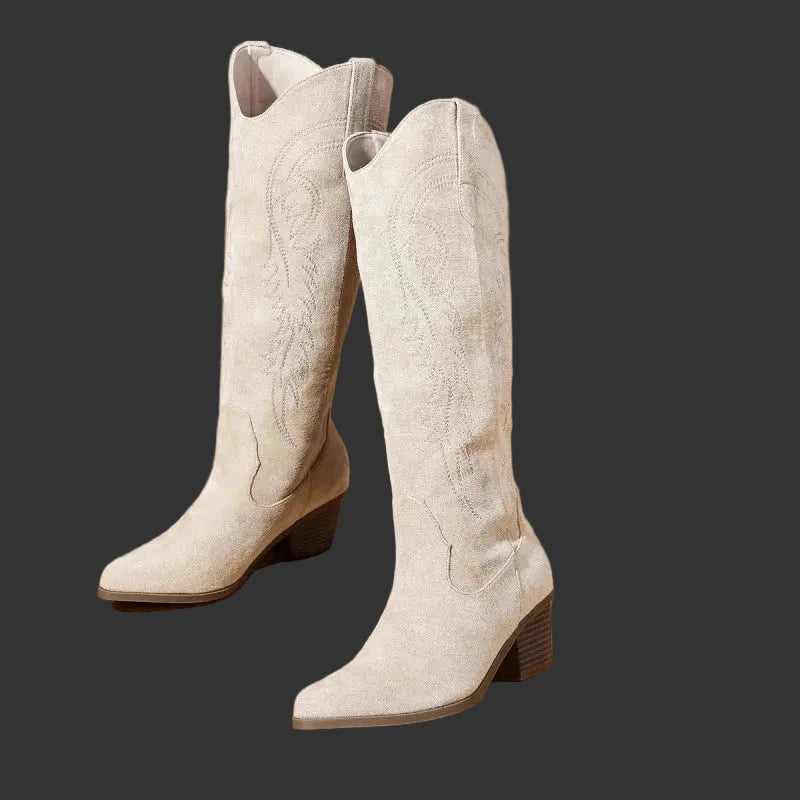 A pair of light beige suede cowboy boots with pointed toes and low block heels, standing on a warm beige background with dried floral decor in the upper left.