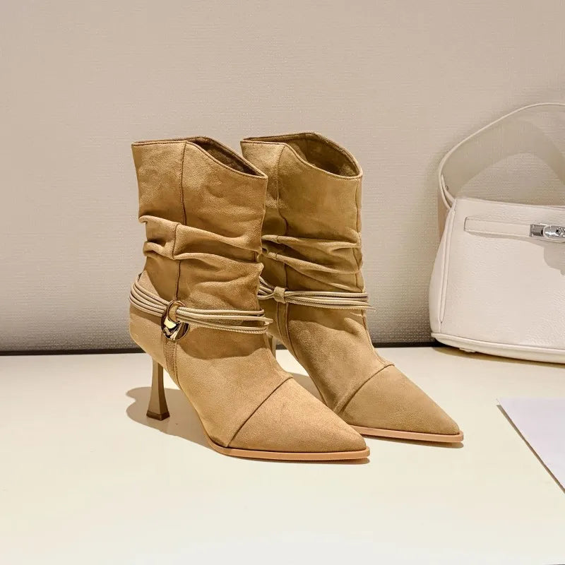A pair of beige suede ankle boots with pointed toes, ruched detailing, and a slim kitten heel, displayed on a light surface next to a white handbag.