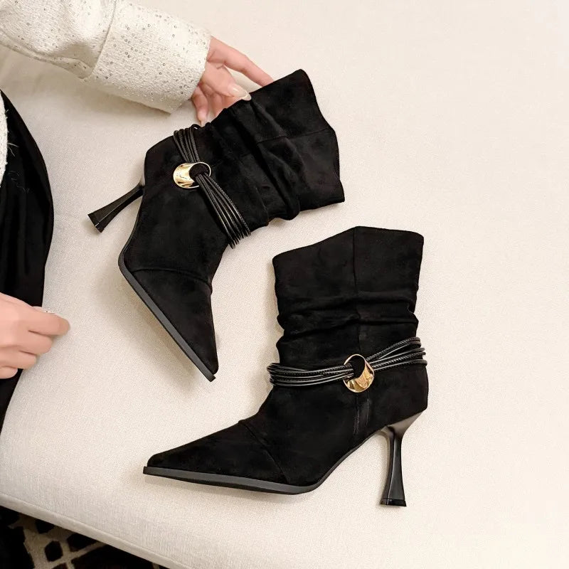 A pair of black suede pointed-toe ankle boots with a high stiletto heel, a gathered elastic strap, and a gold-tone circular accent, shown on a light beige textured surface with a hand holding one boot.