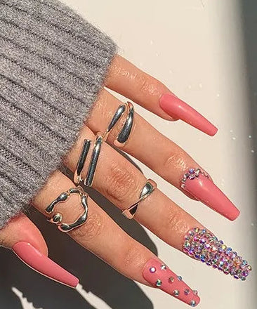 A hand wearing long pink press-on nails with silver rings and glittery nail art, alongside a small inset showing the set of pink press-on nails. A purple label reads '# 1327'.