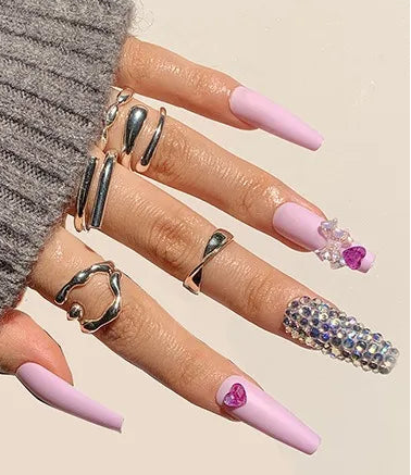 A hand wearing long pastel pink press-on nails with some decorated with rhinestones, shown over a gray ribbed sweater cuff. A small inset at the bottom shows the full set of nail tips, and a purple label reads #1308.