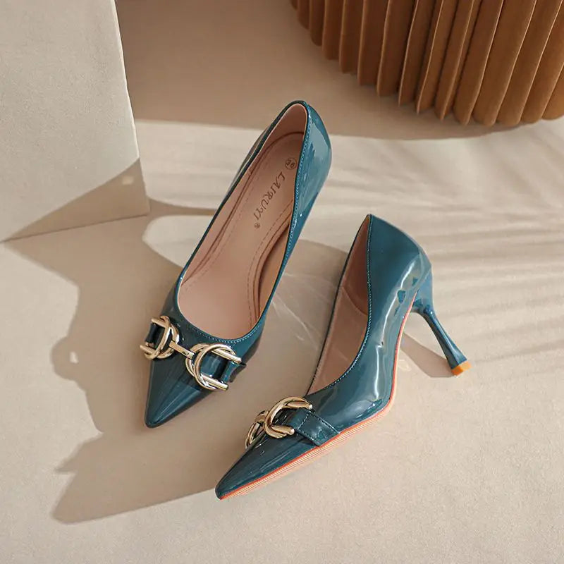 A pair of teal patent leather kitten heels with gold-tone horsebit hardware on the toes, displayed on a beige surface with soft shadows.