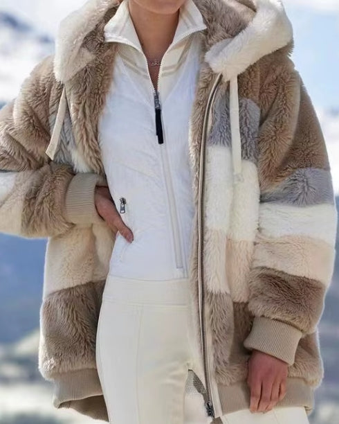 A person wearing a plush hooded jacket with beige, cream, and taupe color-blocked panels over a white zip-front top and white pants, photographed outdoors with a snowy mountain background.