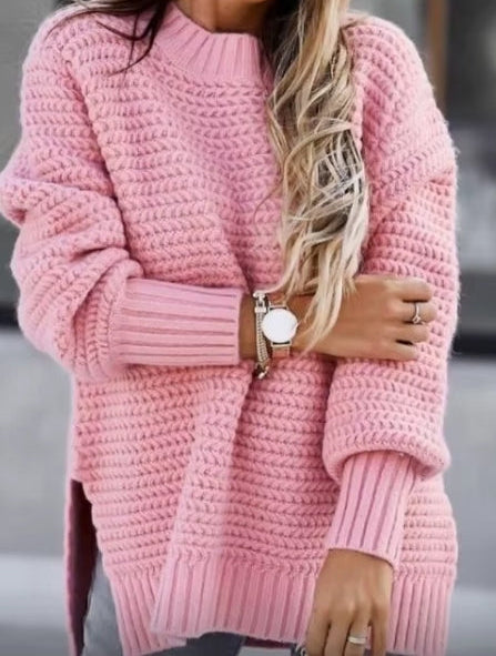 A woman wearing a chunky pink knit sweater, styled with black sunglasses, a gold watch, and rings, set against a blurred gray background.