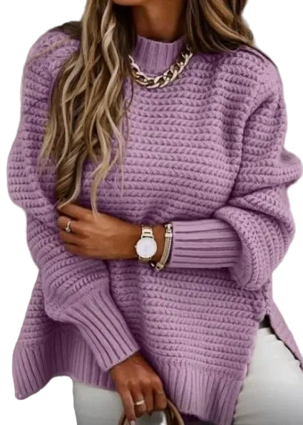 A woman wearing a chunky knit lavender-purple sweater with a mock neck and ribbed cuffs, styled with a chunky gold chain necklace, gold bracelets, and white pants.
