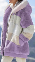 A close-up of a person wearing a fuzzy patchwork jacket in lavender purple and cream, photographed outdoors with a blurred mountain and sky background.