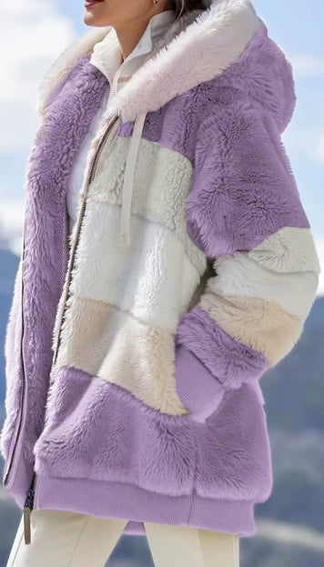 A close-up of a person wearing a fuzzy patchwork jacket in lavender purple and cream, photographed outdoors with a blurred mountain and sky background.